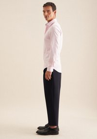 Camicia rosa chiaro a maniche lunghe con colletto, abbinata a pantaloni blu scuro e scarpe nere. La camicia presenta una leggera texture e un orlo curvato.