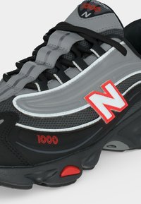 Κοντινό πλάνο γκρι και μαύρου παπουτσιού New Balance 1000 με κόκκινο λογότυπο και λεπτομέρειες, μαύρα κορδόνια και λεπτομέρειες από δίχτυ και συνθετικό υλικό στην επάνω επιφάνεια.
