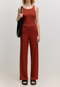 Roestkleurig jumpsuit van ribbelstof, met een aansluitende top en wijde pijpen. Het model draagt een zwarte totebag en zwarte sandalen.