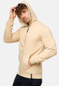 Beige zip-up hoodie med snoremund, der har frontlommer, et logo på brystet og kontrasttekst langs kanten af huen.