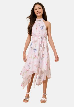 Mädchen in einem ärmellosen, geblümten Neckholderkleid mit asymmetrischem Saum und Taillengürtel, trägt weiße Sandalen und ein Armband, steht vor weißem Hintergrund.