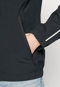 Černá lehká bunda s kapsou na zip a bílým pruhem na rukávu. Elastické manžety a spodní lem zajišťují lepší přiléhavost a pohodlí.