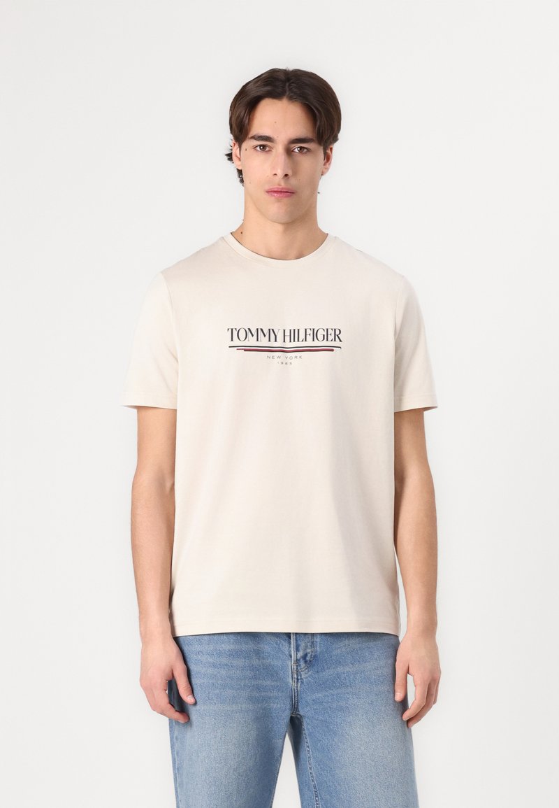 Beige pamut póló kerek nyakkal, melyen a "TOMMY HILFIGER" logó sötétkékkel és pirossal van a mellkason. Világoskék farmernadrággal viselve.