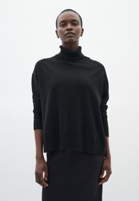 Maglione nero a collo alto con maniche lunghe, vestibilità ampia e polsini a coste. La texture sembra morbida e lavorata a maglia, abbinata a una gonna nera.