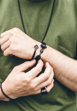 Juoda odinė apyrankė su sudėtingu mazgo raštu, dėvima ant riešo. Rankos pritaiko apyrankę prie žaliai marškiniai.