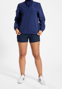 Mörkblå lättviktsjacka med hög krage och dragkedja, kombinerad med mörkblå shorts. Modellen har vita sneakers med svarta detaljer.