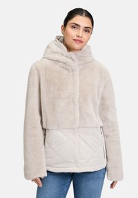 Veste en faux fur beige clair avec un design court, grande capuche et bas matelassé de couleur plus claire, dotée de poches zippées sur les côtés.