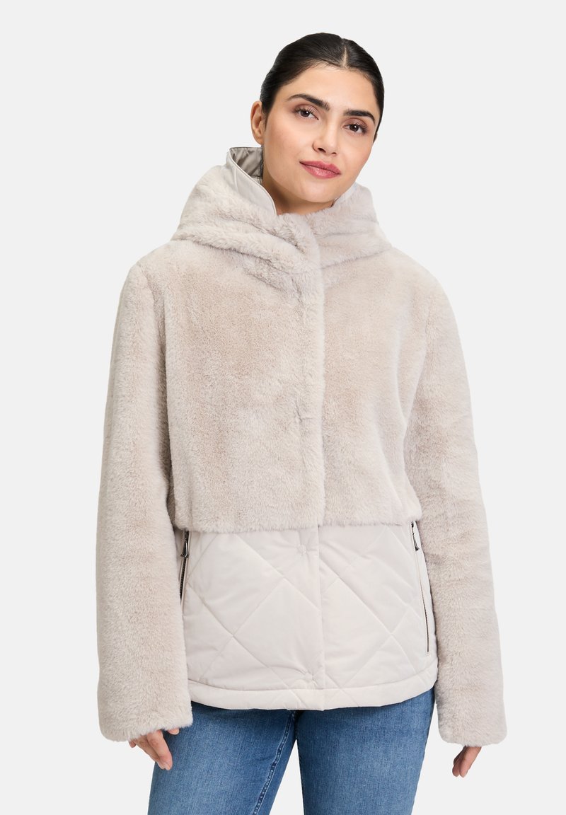 Veste en faux fur beige clair avec un design court, grande capuche et bas matelassé de couleur plus claire, dotée de poches zippées sur les côtés.