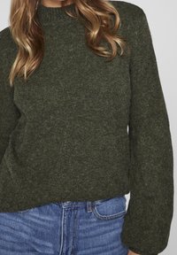 Olivgrön stickad tröja med hög halsringning och långa ärmar, som har en mjuk textur och en avslappnad passform. Bärs över blå jeans.