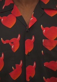 Camicia nera con bottoni e grandi cuori rossi, arricchita da piccole corna di diavolo stampate su tutta la superficie, con due tasche sul petto.