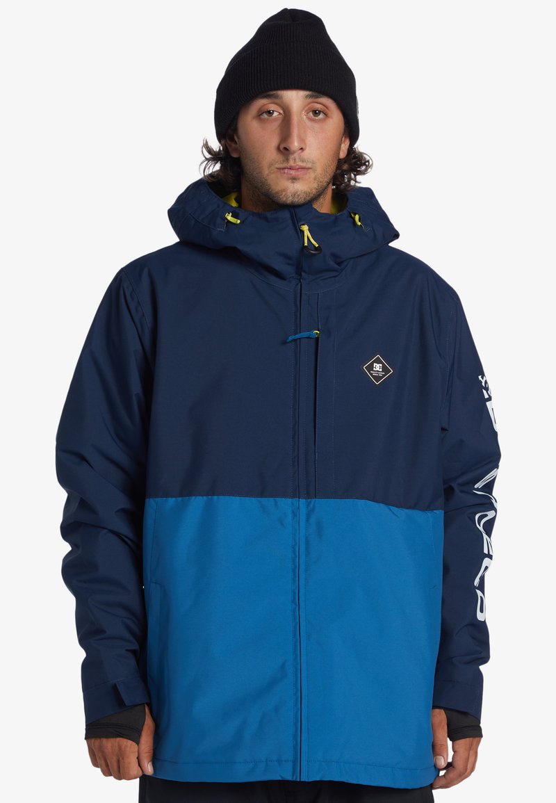 Marinblå och blå tvåfärgad snowboardjacka med huva, gula dragsnören och en logotypapplikation. Strukturerad tyg med elastiska manschetter och fickor.