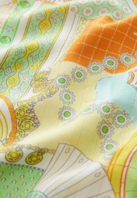 Krāsaina auduma gabals ar ziedu, ģeometriskām un ķēžu rakstām zaļā, oranžā, dzeltenā un zilā krāsā, rādot maigas locītavas un tekstūru.