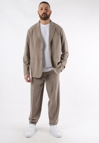 Abito beige con giacca a due bottoni e pantaloni abbinati, realizzato in tessuto morbido. Indossato sopra una maglietta bianca, abbinato a sneakers bianche.