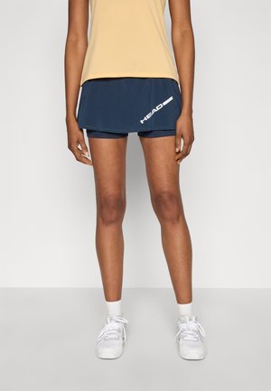 Jupe de sport marine avec shorts intégrés ; conçue en tissu respirant. Présente un détail de logo blanc et est associée à des chaussures de sport blanches.