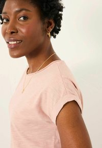 Light pink short-sleeve shirt featuring rolled cuffs, soft texture, round neckline, worn with a delicate gold chain necklace.