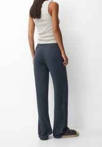 Lange, donkergrijze broek met een losse pasvorm, gemaakt van zacht materiaal. Draagt een beige mouwloos topje en donkere sandalen met een gevlochten zool.