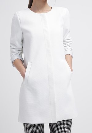 Femme portant un long manteau blanc texturé avec fermeture avant dissimulée et les mains dans les poches, associé à un pantalon pied-de-poule noir et blanc.