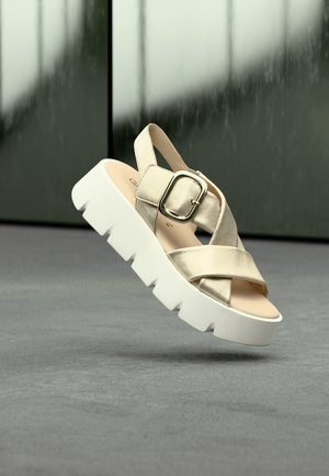 Beige metallic plateauzandalen met gekruiste bandjes, verstelbare gesp en een stevige witte zool, geplaatst op een grijze ondergrond.