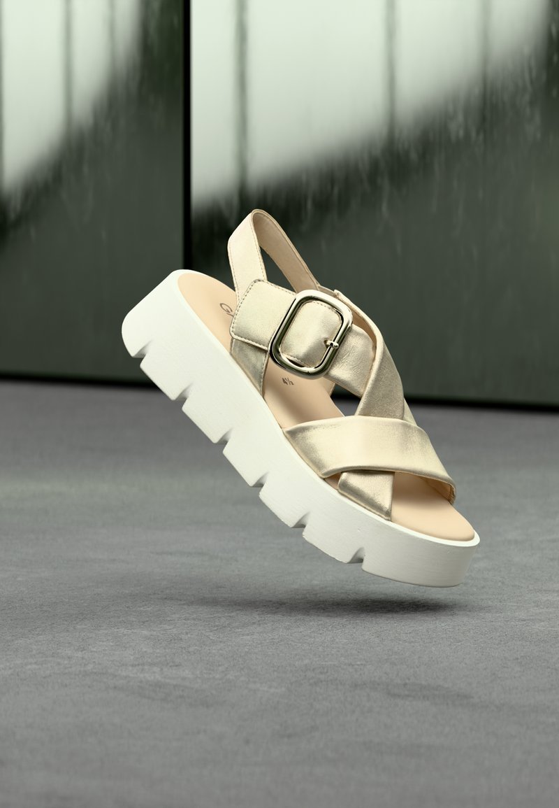 Beige Metallic-Plattform-Sandalen mit gekreuzten Riemen, verstellbarer Schnalle und dicker weißer Sohle, die auf einer grauen Oberfläche liegen.