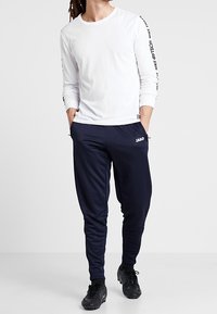 Muž stojící s rukama v kapsách, oblečený v bílém tričku s dlouhým rukávem se textem na rukávech a v námořnických jogger kalhotách s černými teniskami.