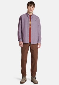 Camisa de botones lila clara superpuesta a una camiseta gráfica roja, combinada con pantalones marrones y botas grises. Diseño minimalista con corte clásico.