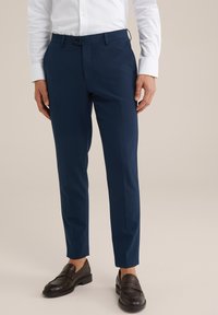 Homme portant un pantalon de costume slim-fit bleu marine, une chemise blanche et des mocassins marron foncé, se tenant contre un fond uni.