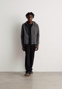 Felpa grigia con zip e cappuccio, t-shirt nera, pantaloni neri e scarpe marroni. Modello in piedi su un pavimento beige contro un muro bianco.