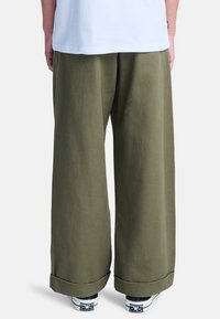 Pantaloni a gamba larga verde oliva realizzati in materiale morbido, con una superficie liscia e orli arrotolati. Indossati con scarpe bianche e una camicia chiara.
