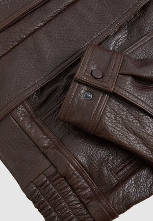 Detail einer dunkelbraunen, strukturierten Lederjacke zeigt genähte Manschette mit Druckknopf und gerafften elastischen Saum.