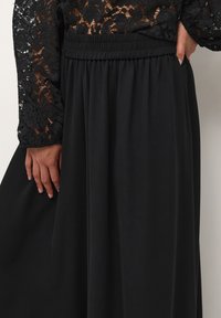 Jupe longue noire en tissu doux, avec une taille élastique et une silhouette fluide. Superposition de dentelle noire détaillée sur les manches.