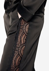 Pantalon en satin noir avec un panneau en dentelle présentant des motifs complexes, un tissu texturé et une finition lisse. Poche latérale visible.