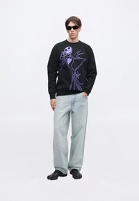 Jovem vestido com um sweat negro com um gráfico de esqueleto roxo, jeans de perna larga azul claro, sapatos negros e óculos de sol pretos, de pé contra um fundo branco.