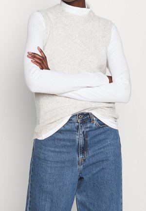 Personne portant un haut blanc à manches longues sous un pull sans manches beige, associé à un jean bleu taille haute, les bras croisés.
