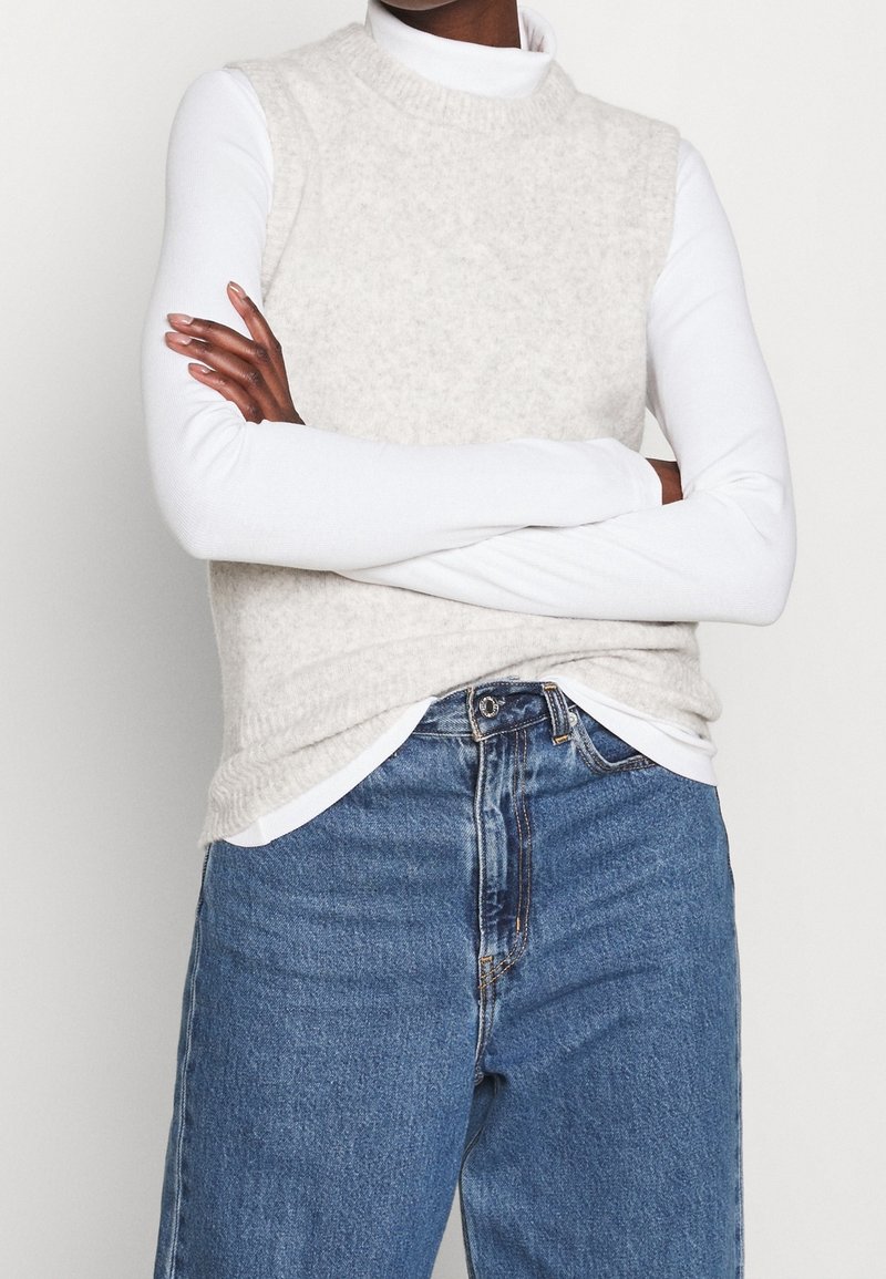 Personne portant un haut blanc à manches longues sous un pull sans manches beige, associé à un jean bleu taille haute, les bras croisés.