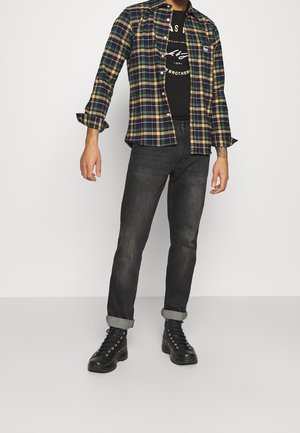 Camisa de franela en patrón de cuadros en azul marino, verde y amarillo, superpuesta sobre una camiseta gráfica negra; combinada con jeans oscuros y botas negras de cordones.