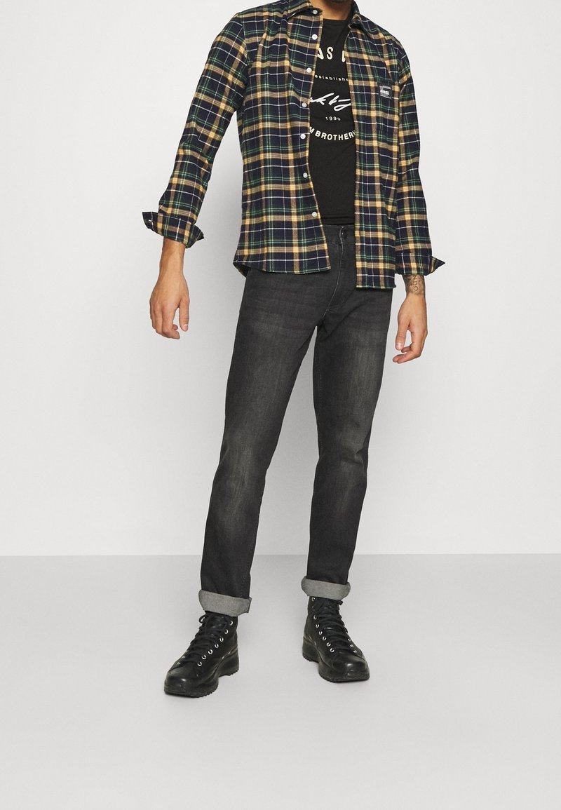 Camisa de franela en patrón de cuadros en azul marino, verde y amarillo, superpuesta sobre una camiseta gráfica negra; combinada con jeans oscuros y botas negras de cordones.