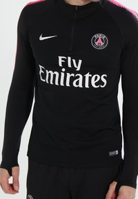 Schwarzes langärmliges Sportoberteil mit einem Viertelreißverschluss, mit pinken Akzenten an den Schultern, "Fly Emirates"-Logo in Weiß und PSG-Emblem.