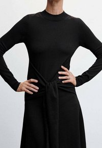 Robe noire en tricot à manches longues, avec un détail noué à la taille. Texture lisse et haut cintré menant à une partie inférieure fluide.