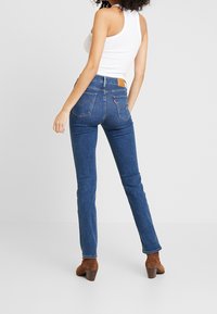 Jeans ajustados de mezclilla en color azul con una etiqueta de cuero en la parte trasera de la cintura. El modelo lleva botas de tacón marrones y una camiseta sin mangas blanca.