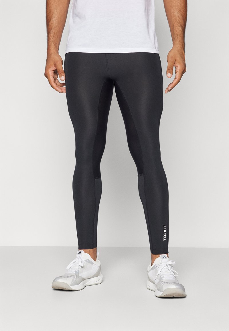 Sorte atletiske leggings lavet af glat, strækbart materiale, med en tætsiddende pasform og et lille hvidt "TECH FIT" logo på den nederste del af benet.