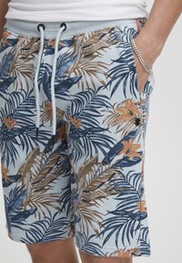 Shorts bleu clair avec un motif de feuilles de palmier tropicales en bleu, marron et orange. Dotés d'une taille élastique et d'un cordon de serrage.