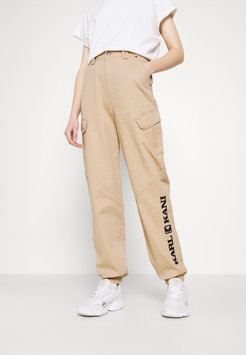 Personne portant un pantalon cargo beige avec un texte noir "KARL KANI" sur la jambe gauche et des baskets blanches, debout avec une main dans la poche.