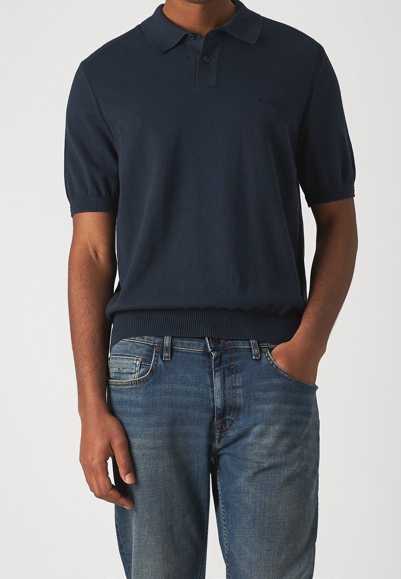 Homme portant un polo à manches courtes bleu marine foncé avec col et un jean bleu, main dans la poche, debout devant un fond uni.