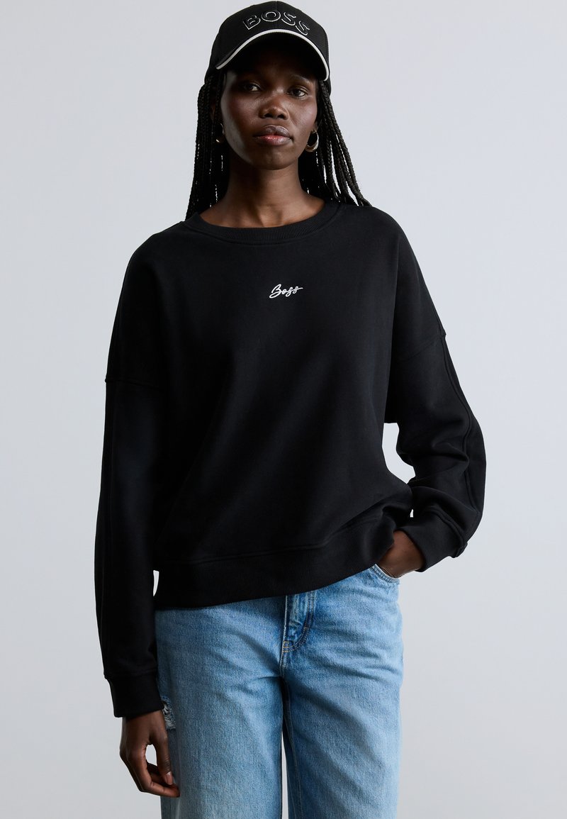 Μαύρο υπερμεγέθες φούτερ με στρογγυλή λαιμόκοψη, διακοσμημένο με λευκή κεντητή επιγραφή "BOSS". Συνδυάζεται με ανοιχτόχρωμο μπλε τζιν με ξεβάμματα και μαύρο καπέλο.
