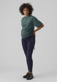 Camiseta premamá de algodón verde oscuro con detalle fruncido lateral y un gráfico de corazón; combinada con leggings azul marino y zapatos negros.