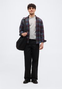 Νέος άνδρας όρθιος, φοράει καρό πουκάμισο πάνω από γκρι μπλουζάκι, μαύρο παντελόνι, μαύρα παπούτσια και κρατά μια μεγάλη μαύρη τσάντα ώμου.