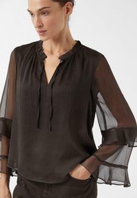 Blouse noire transparente avec col à volants, décolleté en V et manches longues ornées de volants superposés. Texture lisse avec une coupe décontractée.