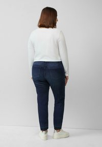 Dunkelblaue Jeans mit ausgewaschenen Akzenten, vier Taschen und slim fit, kombiniert mit einem weißen Langarmoberteil und weißen Sneakers.
