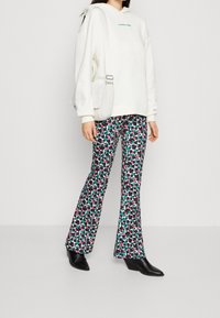Felpa bianca con cappuccio e testo verde, abbinata a pantaloni a zampa d'elefante con un motivo floreale multicolore su sfondo nero. Stivaletti neri.