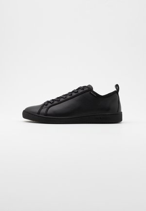 Baskets basses en cuir noir avec lacets noirs, bout arrondi et languette au talon sur fond gris clair.